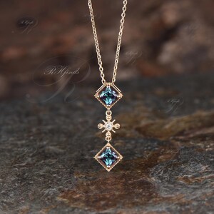 Op de afbeelding: Een gouden ketting met een hanger met drie vierkante edelstenen. De bovenste en onderste edelstenen zijn diepblauwgroen, terwijl de middelste edelsteen helder wit is. De hanger is versierd met ingewikkelde gouden details.