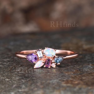 May include: A rose gold ring featuring a cluster of multi-shaped gemstones, including two white opals, two purple pear and baguette-cut stones, and a blue baguette-cut stone. The band is thin and polished, with the text RHfinds visible in the background.