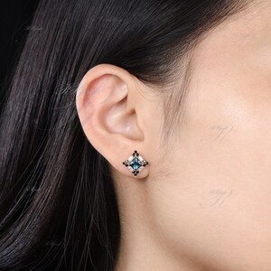 May include: A square-shaped stud earring with a teal center stone, surrounded by black and white accent stones. The earring is set in a gold-colored metal and is displayed on an ear.