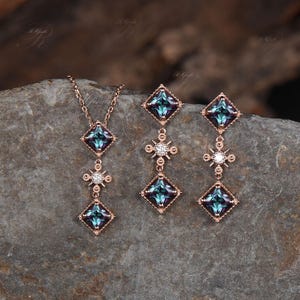 Op de afbeelding: Een driedelige sieradenset bestaande uit een ketting en oorbellen. De ketting en oorbellen zijn rose goud van kleur en hebben een vierkante blauwe edelsteen met een kleinere diamantvormige edelsteen eronder. De oorbellen hebben een kleinere diamantvormige edelsteen bovenaan.