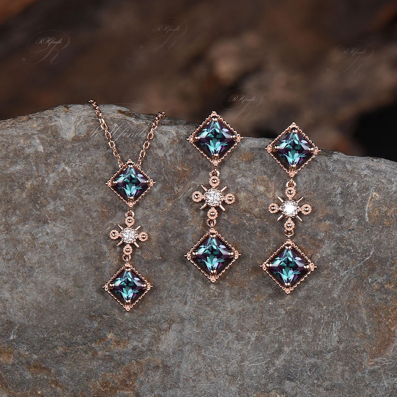 Op de afbeelding: Een driedelige sieradenset bestaande uit een ketting en oorbellen. De ketting en oorbellen zijn rose goud van kleur en hebben een vierkante blauwe edelsteen met een kleinere diamantvormige edelsteen eronder. De oorbellen hebben een kleinere diamantvormige edelsteen bovenaan.