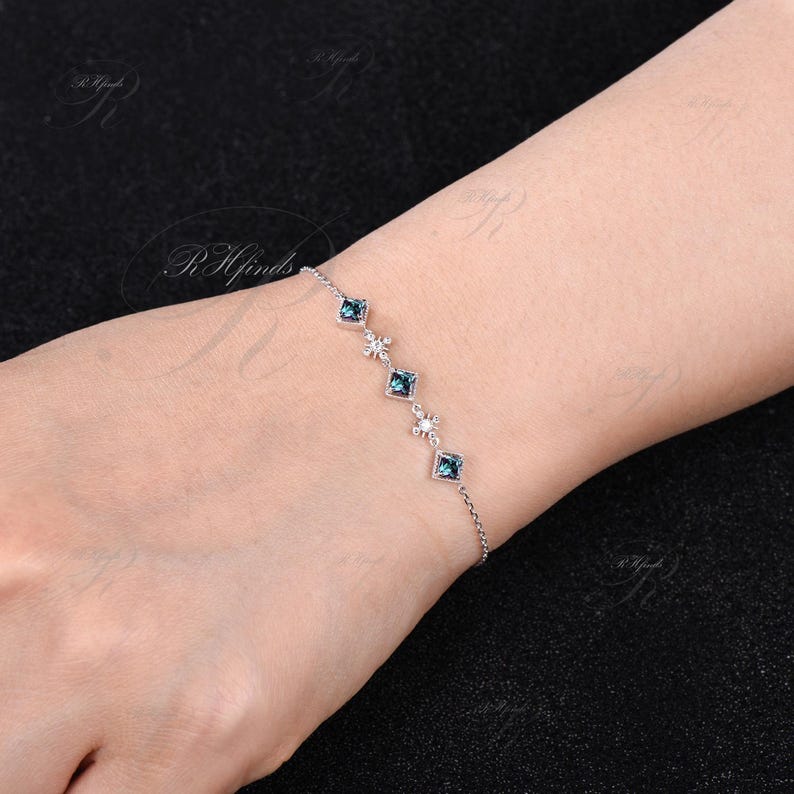 Op de afbeelding: Een delicate zilveren kettingarmband met vijf vierkante blauwe edelstenen en kleine diamant-achtige accenten.