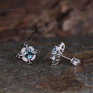 May include: A pair of stud earrings featuring a square, teal-colored center stone. The earrings are surrounded by a halo of black and white stones, set in a silver-colored metal. The earrings are displayed on a dark, textured surface.