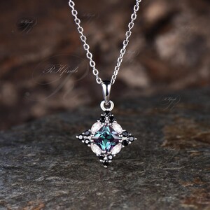 May include: A silver pendant necklace featuring a square-shaped pendant with a central teal-colored stone. The pendant is adorned with black and white accent stones, suspended from a delicate chain. The jewelry is displayed on a textured, dark gray surface.