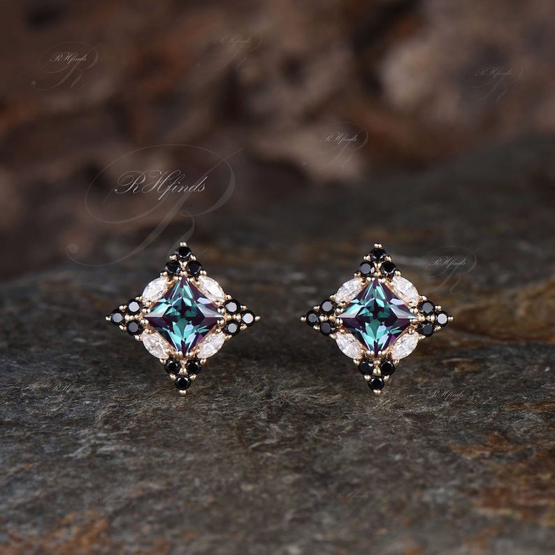 May include: A pair of gold-toned stud earrings. Each earring features a square, teal-colored center stone, surrounded by black and white accent stones in a geometric design. The earrings are set on a dark, textured surface.
