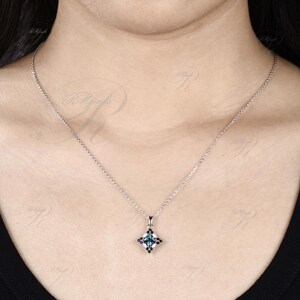 May include: A silver necklace with a square pendant featuring a teal-colored gemstone at the center, surrounded by smaller black stones. The pendant hangs from a delicate chain, creating a classic and elegant look. The necklace is displayed against a neutral background.