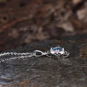 May include: A silver pendant necklace featuring a central, round, teal-colored gemstone. The gemstone is surrounded by smaller, black accent stones. The necklace is displayed on a dark, textured surface.