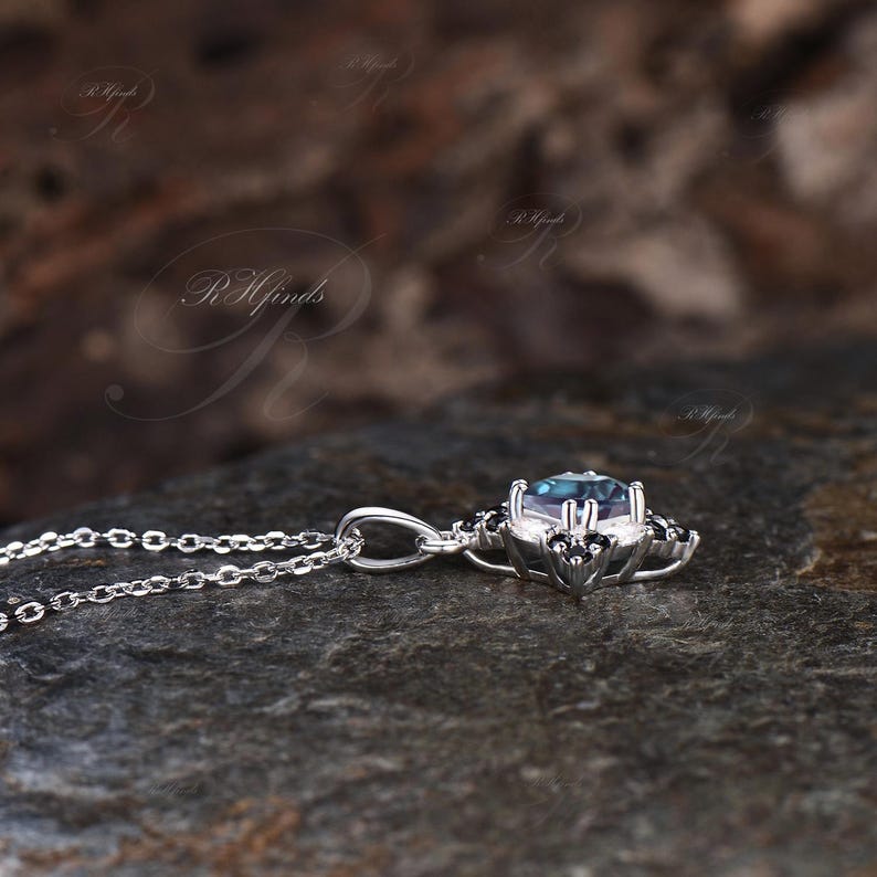 May include: A silver pendant necklace featuring a central, round, teal-colored gemstone. The gemstone is surrounded by smaller, black accent stones. The necklace is displayed on a dark, textured surface.
