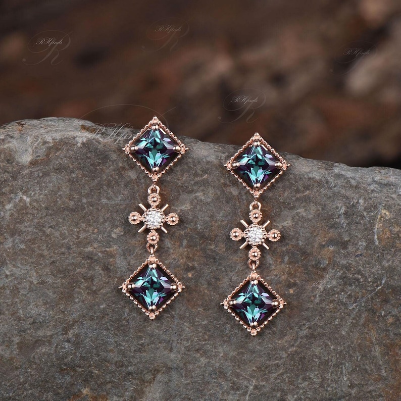Op de afbeelding: Een paar oorbellen van ros&eacute;goud met een vierkante blauwe edelsteen en een kleinere diamantvormige edelsteen in het midden van een kruisvormig ontwerp.