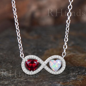 May include: A silver-toned infinity pendant necklace featuring a heart-shaped red gemstone and a heart-shaped white iridescent gemstone, both surrounded by small clear stones. The metal chain is composed of oval links. The text RHfonds is visible.