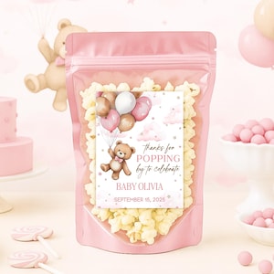 Op de afbeelding: Roze popcornzak met een label met de tekst "bedankt voor het langskomen om te vieren" met een teddybeer met ballonnen. De zak is gevuld met popcorn en is omgeven door roze decoraties, waaronder een cake en snoep.