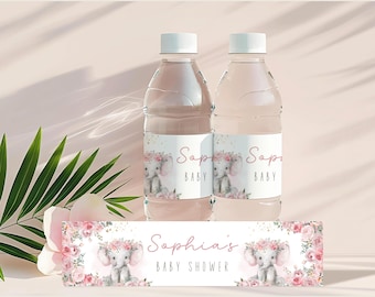 Può includere: Due bottiglie d'acqua trasparenti con tappi bianchi ed etichette raffiguranti un elefantino con una corona floreale. Le etichette riportano "Sophia's BABY" e sono circondate da fiori rosa. Un banner abbinato è di fronte.