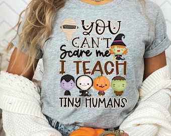 May include: A gray t-shirt with the text "You Can't Scare Me I Teach Tiny Humans" and Halloween-themed graphics. The design includes a mummy, witch, vampire, skeleton, pumpkin, and Frankenstein's monster.