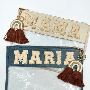 Può includere: Due pochette trasparenti con cerniera, una color crema e una blu denim, ciascuna con un nome in lettere beige strutturate. Entrambe hanno un ciondolo a forma di arcobaleno con nappe marroni. La pochette color crema dice "MAMA" e quella blu dice "MARIA".