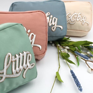 Pode incluir: Quatro bolsas cosméticas retangulares em cores pastel: verde menta, coral, azul empoeirado e creme. Cada bolsa tem a palavra "Little" ou "Big" bordada em escrita branca. Um ramo de lavanda e vegetação está em primeiro plano.