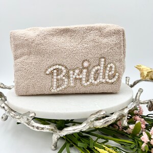 Könnte beinhalten: Eine beige Kosmetiktasche aus Frottee mit dem Wort "Bride" in Perlen und Goldfaden gestickt. Die Tasche steht auf einem weißen Marmorsockel, umgeben von Grün und einer dekorativen silbernen Metallbasis.