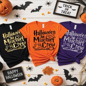 May include: Three short-sleeve t-shirts in black, orange, and purple, each featuring the text "Halloween Mischief Crew" with graphics of a ghost, a broom, a spider, and a bat. The black shirt has gold text, the orange has black, and the purple has white.