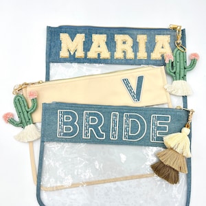 Op de afbeelding: Drie denim ritsvakken met doorzichtige plastic ramen. Het bovenste vak heeft de naam "MARIA" geborduurd in gouden letters. Het middelste vak heeft een beige stof met een grote "V" geborduurd in blauw. Het onderste vak heeft het woord "BRIDE" geborduurd in wit. Elk vak heeft een kwastje.