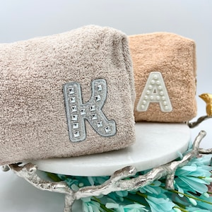 May include: Two beige terry cloth cosmetic bags. One bag features a silver letter "K" embellished with silver studs. The other bag has a white letter "A" made of pearls. The bags are displayed on a white marble stand.