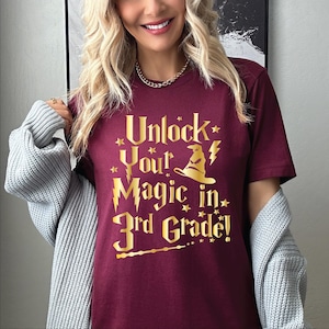 May include: A maroon t-shirt with gold text that reads "Unlock Your Magic in 3rd Grade!" The design includes a wizard hat, lightning bolt, stars, and a wand. The shirt is part of a back-to-school theme.