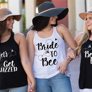 Könnte beinhalten: Drei Frauen tragen Tanktops mit Texten und Designs zum Thema Bienen. Ein Tanktop ist weiß mit "Bride to Bee", ein anderes ist schwarz mit "Bee Hive", und das dritte ist schwarz mit "Let's Get Buzzed". Sie tragen Hüte.
