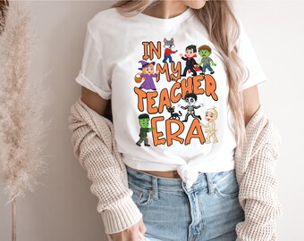May include: White t-shirt with the text "IN MY TEACHER ERA" in orange, featuring cartoon Halloween characters. The design includes a witch, vampire, zombie, mummy, and other spooky figures. The shirt is worn by a person.