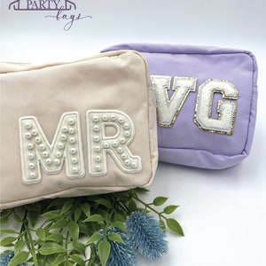 May include: Two zippered cosmetic bags with pearl-embellished letters. The bag on the left is beige with the letters "MR" and the bag on the right is lavender with the letters "VG".