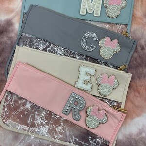 Op de afbeelding: Vier pastelkleurige etuis met ritssluiting en transparante voorkant. Elke etui heeft een letter en een Minnie Mouse-hoofdontwerp. De letters zijn versierd met parels, glitter en studs. De etuis zijn in tinten blauw, grijs, crème en roze.