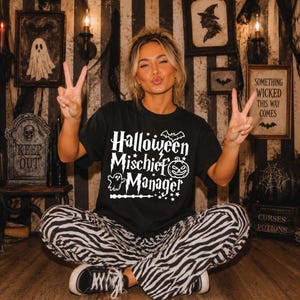 May include: A black short-sleeve t-shirt featuring white graphic text that reads Halloween Mischief Manager, accompanied by illustrations of a bat, a jack-o'-lantern, a ghost, a magic wand, and stars, paired with black and white zebra print pants.