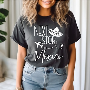Könnte beinhalten: Dunkelgraues T-Shirt mit dem weißen Text "NEXT STOP Mexico". Das Design zeigt einen Sombrero, ein Flugzeug und eine gepunktete Linie. Das T-Shirt ist ein lässiger Stil.