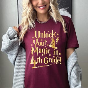 May include: A maroon t-shirt with gold text that reads "Unlock Your Magic in 4th Grade!" The design includes a wizard hat, lightning bolt, stars, and a wand. The shirt is short-sleeved.