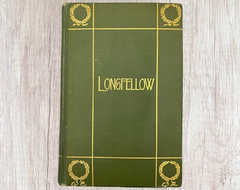 May include: A hardcover book with a dark green cover and gold lettering that reads "LONGFELLOW." The cover features gold laurel wreath designs in each corner and gold lines. The book is set against a light wood grain background.