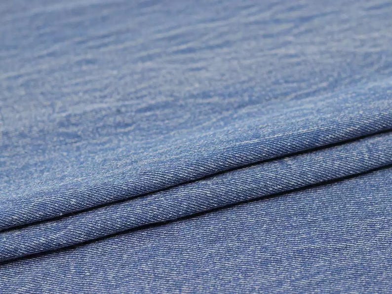 Puede incluir: Primer plano de una tela vaquera azul con un sutil patr&oacute;n texturizado. La tela est&aacute; doblada por la mitad, mostrando el tejido y la textura.