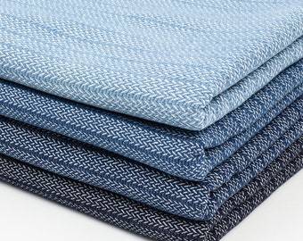 Op de afbeelding: Stapel van vier blauwe en witte stofstalen met een herringbone-patroon. De stalen zijn gevouwen en op elkaar gelegd, met de lichtste blauwe staal bovenop en de donkerste blauwe staal onderaan.