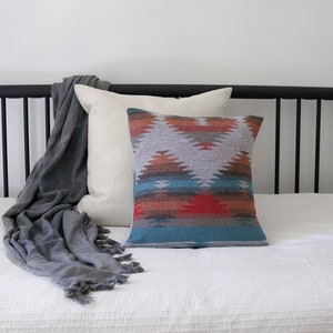 Puede incluir: Una cama blanca con una cabecera de metal negro y una manta gris. Hay dos almohadas en la cama, una blanca y otra con un patrón geométrico colorido.