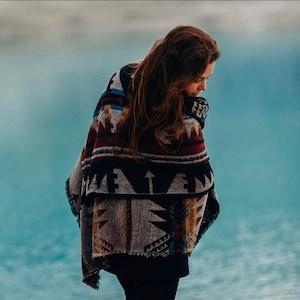 May include: A person wearing a colorful patterned blanket with black, white, red, and blue geometric designs. The blanket is wrapped around the person's shoulders and is fringed at the edges.