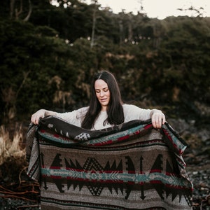 May include: A woman with long dark hair stands in a forest setting, holding a brown and grey patterned blanket with red and blue accents. The blanket has a geometric design.
