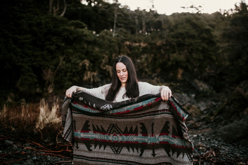 May include: A woman with long dark hair stands in a forest setting, holding a brown and grey patterned blanket with red and blue accents. The blanket has a geometric design.