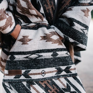 May include: A black, brown, and white patterned poncho with a leather label. The poncho has a geometric design with a fringe border.