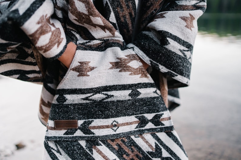 May include: A black, brown, and white patterned poncho with a leather label. The poncho has a geometric design with a fringe border.