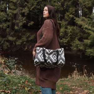 Peut inclure: Une femme portant un pull marron et un jean bleu porte un sac fourre-tout à motif géométrique noir et blanc avec des bretelles noires. Le sac est porté sur son épaule et elle marche dans un environnement forestier.