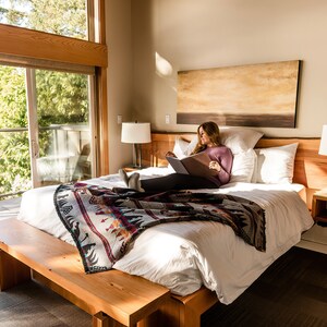 May include: A woman relaxes in a bed with white linens and a colorful patterned throw blanket. The bed is next to a large window with a view of a balcony and trees. A lamp is on the nightstand next to the bed.