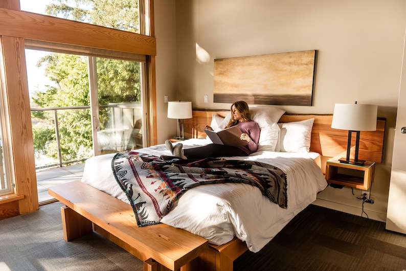 May include: A woman relaxes in a bed with white linens and a colorful patterned throw blanket. The bed is next to a large window with a view of a balcony and trees. A lamp is on the nightstand next to the bed.