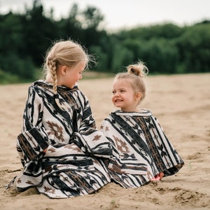 May include: Two young girls wearing black and white patterned ponchos with brown accents. The girls are sitting on a sandy beach and smiling at each other.