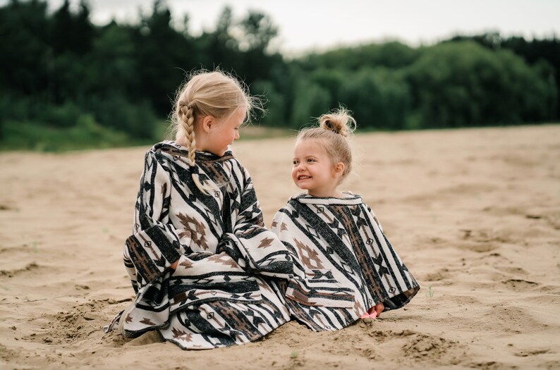 May include: Two young girls wearing black and white patterned ponchos with brown accents. The girls are sitting on a sandy beach and smiling at each other.