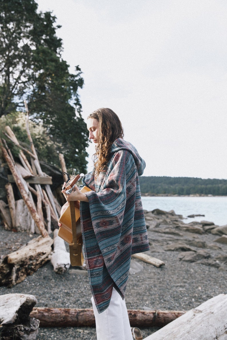 May include: A person wearing a blue and red patterned poncho plays an acoustic guitar on a rocky beach.