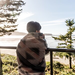 May include: A person wrapped in a colorful blanket stands on a balcony overlooking a beach and ocean. The person is looking out at the view.