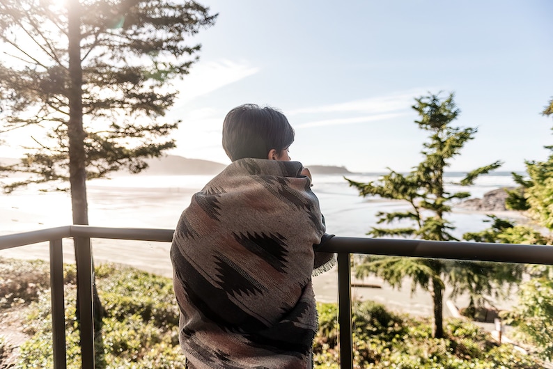 May include: A person wrapped in a colorful blanket stands on a balcony overlooking a beach and ocean. The person is looking out at the view.