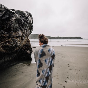 May include: A person wearing a blue and brown patterned blanket walks on a sandy beach with a large rock in the background.