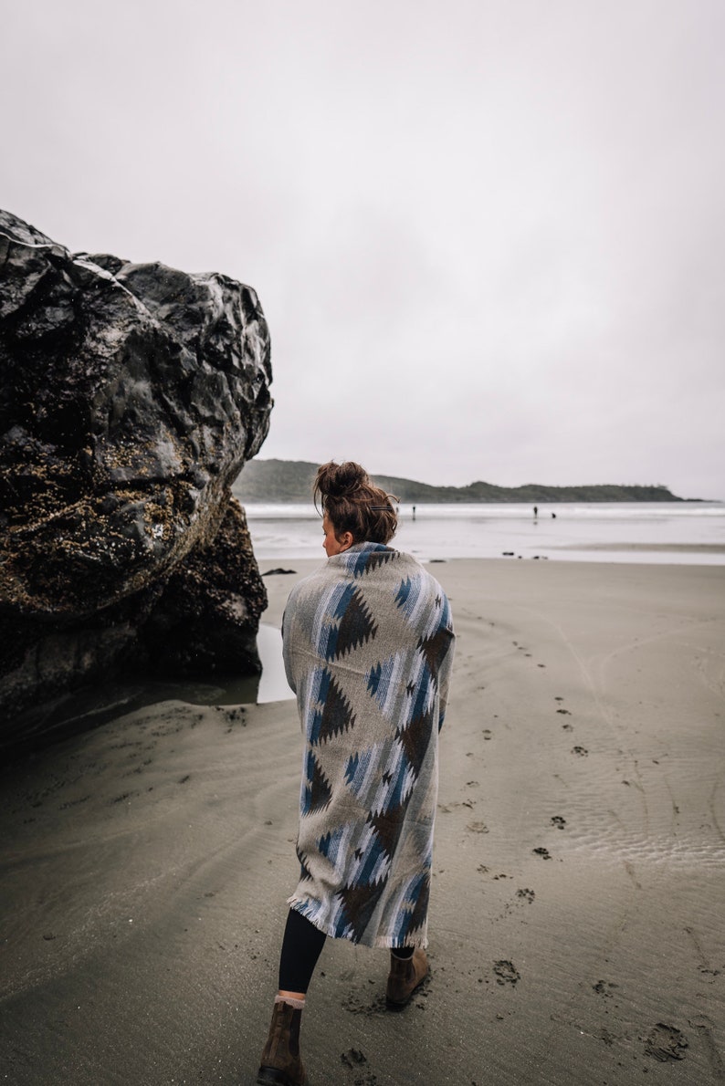 May include: A person wearing a blue and brown patterned blanket walks on a sandy beach with a large rock in the background.